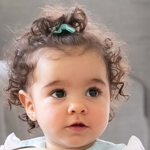 Adorable black haired baby girl in pale blue clothes sitting on floor at home, looking away, playing soccer ball. Kid at home and childhood concept